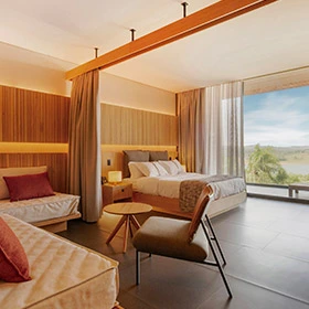 linda vista do quarto amplo de hóspede do resort, um dos quartos incríveis oferecidos pelo local; o ambiente oferece cama de casal e um sofá em L com poltrona para momentos de leitura e descontração, além de uma linda vista para o lago e natureza